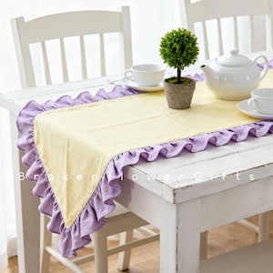 May include: A yellow table runner with lavender frilled edges on a white wooden table. A small potted topiary, white teapot, and teacups are arranged on the table. The table is set for tea.