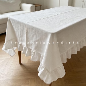 May include: A white linen tablecloth with ruffled edges covers a wooden table. The tablecloth has a simple design and is draped over the table. The text "Brokenflower Gifts" is printed on the tablecloth.