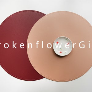 May include: Two round leather placemats, one burgundy and one light peach, with a small white bowl in the center of the peach placemat. The text "brokenflowerGift" is printed in white across the center of the image.