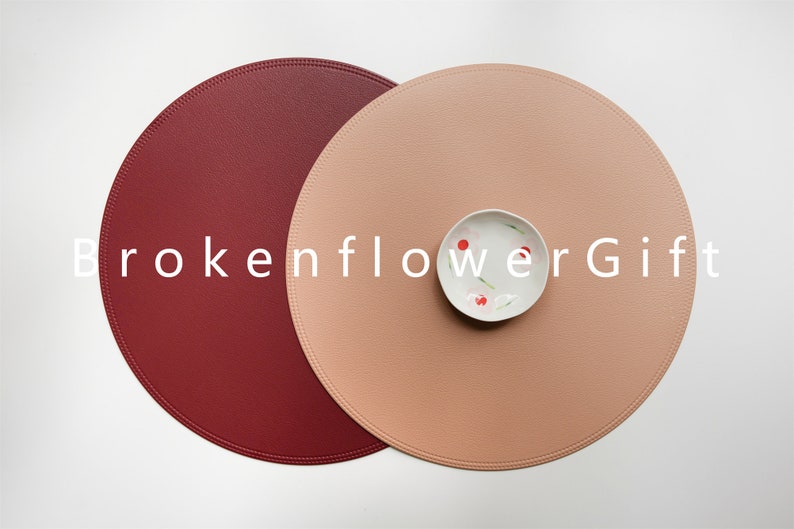 May include: Two round leather placemats, one burgundy and one light peach, with a small white bowl in the center of the peach placemat. The text "brokenflowerGift" is printed in white across the center of the image.