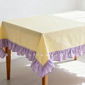 Puede incluir: Un mantel rectangular amarillo con un ribete de volantes lavanda. El mantel está colocado sobre una mesa de madera con patas de color claro. El texto "Brokenflower Gifts" es visible en el mantel.