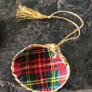 May include: A decorative ornament featuring a shell shape with a gold trim. The shell is covered in a red, green, yellow, and white plaid pattern. A gold tassel and string are attached for hanging.