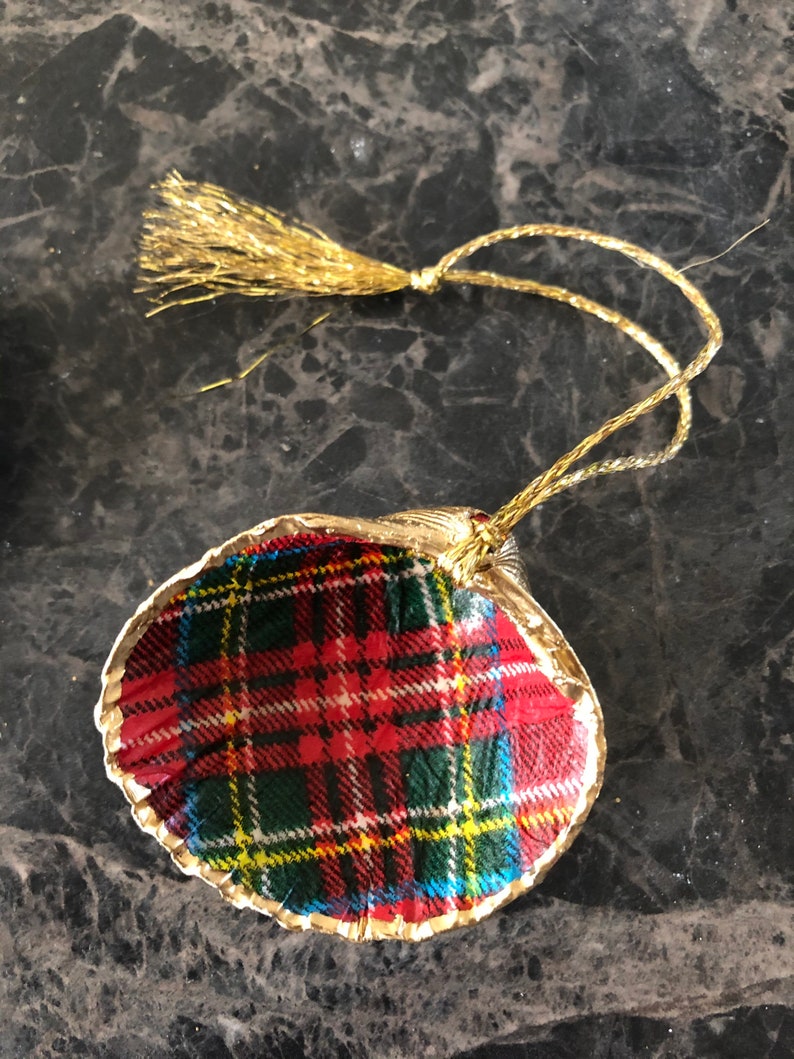 May include: A decorative ornament featuring a shell shape with a gold trim. The shell is covered in a red, green, yellow, and white plaid pattern. A gold tassel and string are attached for hanging.