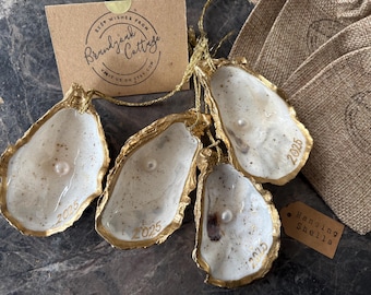 Puede incluir: Cuatro adornos de concha de ostra con bordes pintados en oro, cada uno con una perla blanca en el centro y el año 2025 inscrito en dorado. Las conchas están unidas por un cordón dorado e incluyen una tarjeta de marca y una etiqueta con el texto Hanging Shells.