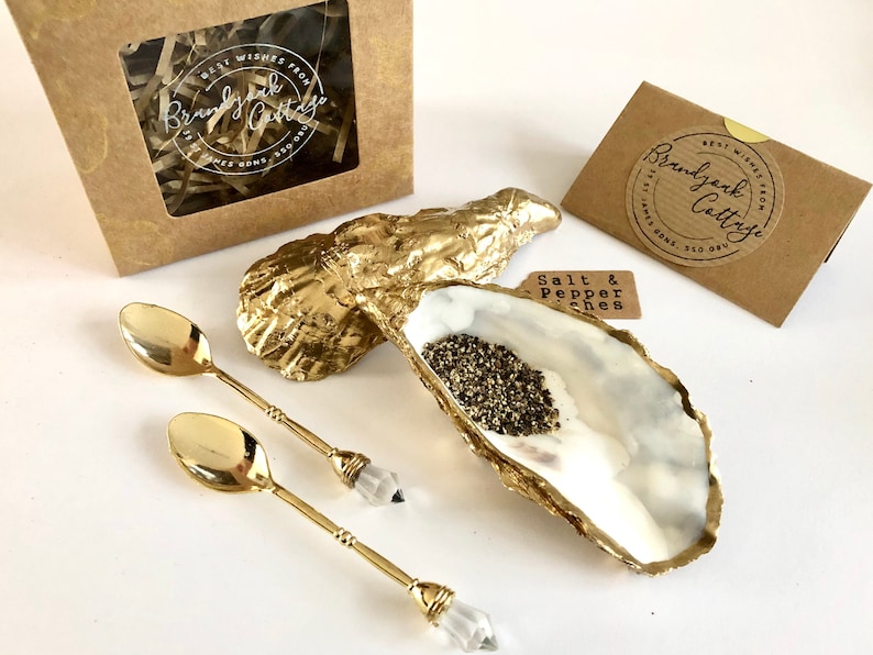 Op de afbeelding: Gouden oesterschelp zout- en peperstrooiers met twee gouden lepels met heldere kristallen handvatten. De set wordt geleverd in een bruine doos met een label waarop staat "Beste wensen van Brandy Oak Cottage".