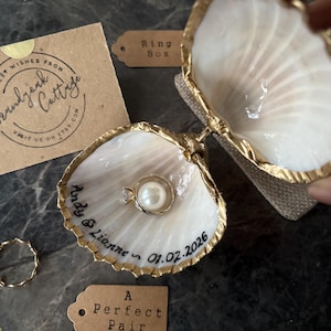 May include: A seashell ring box with gold trim, holding a pearl ring. The box is open, revealing the ring inside. A small gold ring and a tag that says "A Perfect Pair" are also visible. The box is personalized with a name and date.