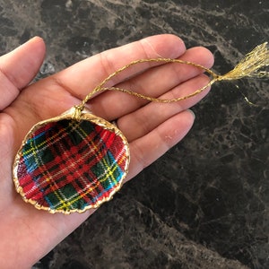 May include: A handcrafted Christmas ornament shaped like a seashell. The ornament features a red, green, and yellow plaid pattern inside a gold-painted shell. A gold string is attached for hanging.