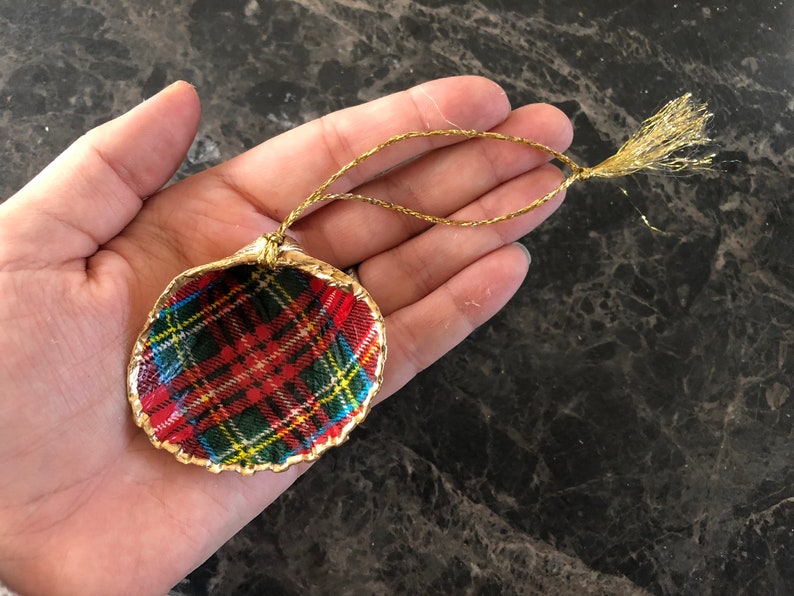 May include: A handcrafted Christmas ornament shaped like a seashell. The ornament features a red, green, and yellow plaid pattern inside a gold-painted shell. A gold string is attached for hanging.