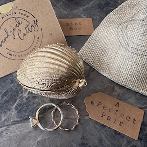 Puede incluir: Una caja de anillos dorada en forma de concha con dos anillos dorados, uno con una piedra preciosa transparente. Tambi&eacute;n hay una peque&ntilde;a bolsa de arpillera y una etiqueta que dice "A Perfect Pair".