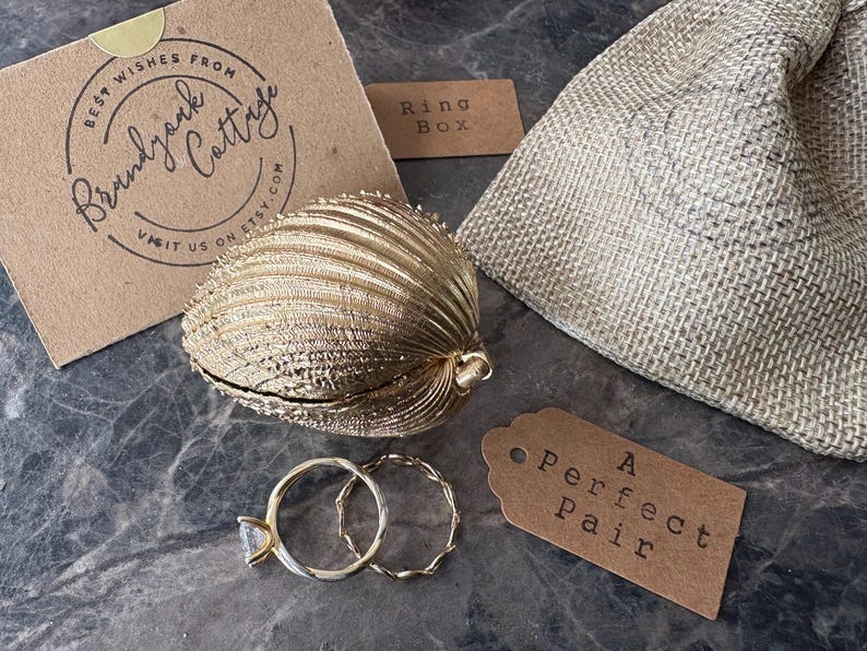 Puede incluir: Una caja de anillos dorada en forma de concha con dos anillos dorados, uno con una piedra preciosa transparente. Tambi&eacute;n hay una peque&ntilde;a bolsa de arpillera y una etiqueta que dice "A Perfect Pair".