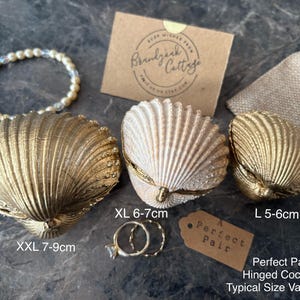 Puede incluir: Cajas de concha con bisagras de varios tama&ntilde;os, de 5-9 cm, en dorado y crema. La imagen incluye una pulsera de perlas y dos anillos. Una etiqueta dice "Perfect Pair".