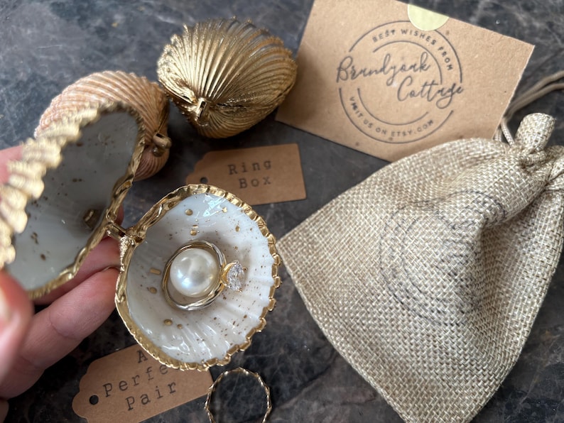 Puede incluir: Un joyero de concha con ribete dorado, que contiene un anillo con una perla y un peque&ntilde;o diamante. La caja est&aacute; abierta, revelando el anillo en su interior. Tambi&eacute;n hay otras conchas y una peque&ntilde;a bolsa de yute.