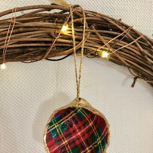 May include: A handcrafted Christmas ornament made from a seashell, featuring a red, green, and yellow plaid pattern. The shell is trimmed with gold paint and hangs from a gold cord. The ornament is set against a rustic wreath.