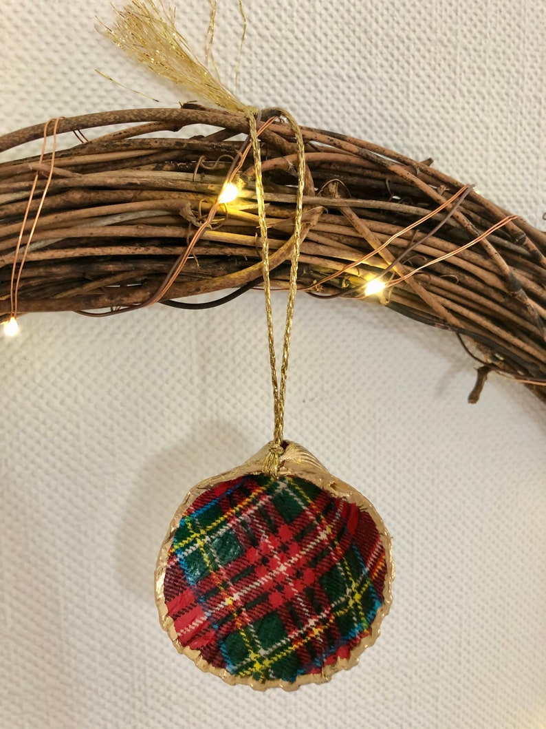 May include: A handcrafted Christmas ornament made from a seashell, featuring a red, green, and yellow plaid pattern. The shell is trimmed with gold paint and hangs from a gold cord. The ornament is set against a rustic wreath.