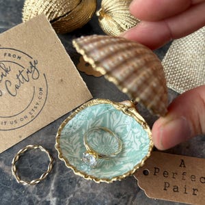 May include: A seashell ring holder with a gold trim and a light blue floral pattern, holding a gold ring with a clear stone. A second gold ring and a small tag that says "A Perfect Pair" are also visible.