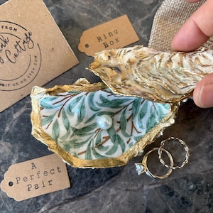 以下が含まれることがあります： 緑と白の花柄の裏地が付いた金色の牡蠣の貝殻のリングボックス。箱が開いており、中に金色のリングが2つ入っています。箱は灰色の表面の上に置かれています。茶色のタグに「A Perfect Pair」の文字があります。