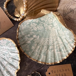 以下が含まれることがあります： 金色の縁取りが施された貝殻の装飾品皿。内側は、水色と白の花と鳥の模様です。パールネックレスと「Trinket Dish」と書かれたタグも表示されています。