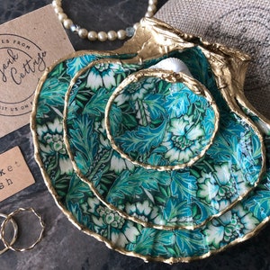 May include: Three nested trinket dishes made from seashells, painted with a green and white floral pattern and accented with gold trim. The dishes are arranged in a stack, with the largest dish on the bottom and the smallest dish on top. The dishes are on a gray surface.