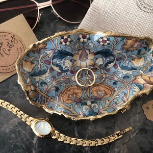 May include: A blue and gold trinket dish with a floral pattern. The dish is made of resin and has a gold rim. The dish is sitting on a gray surface with a gold watch and sunglasses.