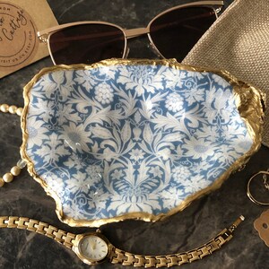 May include: A blue and white ceramic trinket dish with a gold rim. The dish is shaped like a seashell and has a floral pattern. The dish is sitting on a dark surface with a gold watch, a pearl necklace, and two gold rings.