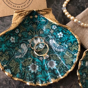 May include: A teal blue and gold seashell ring dish with a floral pattern and two gold rings inside. The dish is made of resin and has a gold rim.