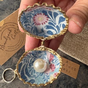 May include: A hinged seashell ornament with a gold-painted rim, featuring an interior pattern of blue foliage and pink flowers. A single white pearl is attached to the bottom shell, and a silver-toned metal ring is connected to the exterior hinge.