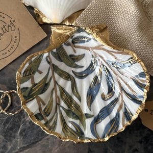 May include: A decorative seashell trinket dish with a gold trim and a botanical pattern in green and blue. Two gold rings rest inside. A small tag reads "Trinke Dish". A card with text is in the background.
