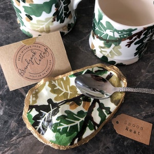 May include: A white ceramic spoon rest with a gold rim and a green and gold leaf pattern. The spoon rest is sitting on a grey surface with a silver spoon resting inside. A brown tag with the text "Spoon Rest" is in the lower right corner of the image.