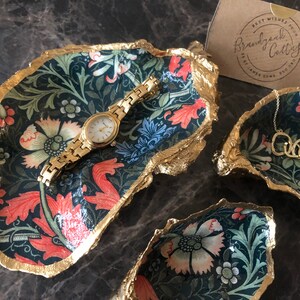 Pu&ograve; includere: Piatti decorativi per bigiotteria con un motivo floreale blu navy, rosso e verde, bordati d'oro. Un orologio d'oro &egrave; appoggiato in un piatto e una collana d'oro in un altro. I piatti sono disposti su una superficie scura.