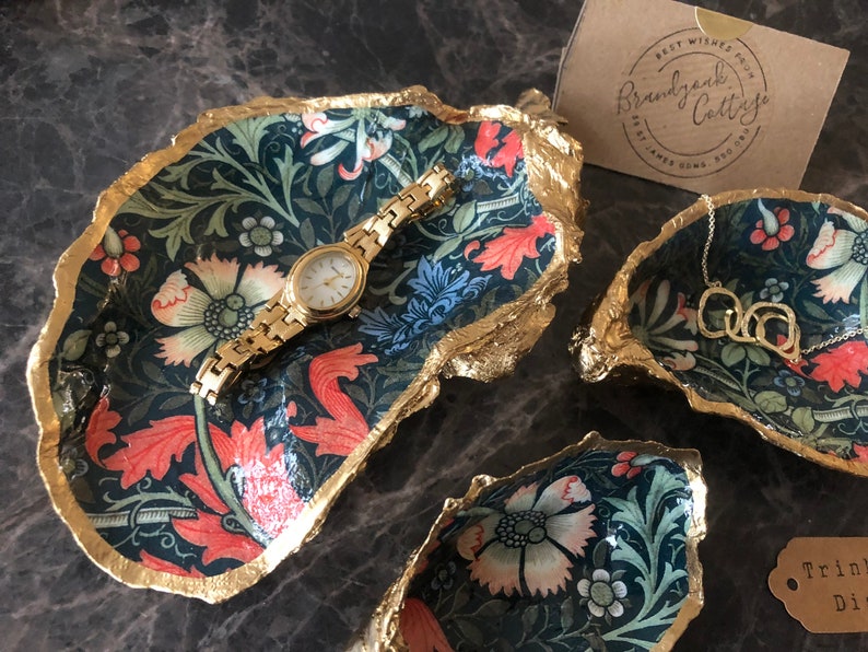 Pu&ograve; includere: Piatti decorativi per bigiotteria con un motivo floreale blu navy, rosso e verde, bordati d'oro. Un orologio d'oro &egrave; appoggiato in un piatto e una collana d'oro in un altro. I piatti sono disposti su una superficie scura.