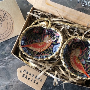 May include: Two gold-painted, hanging shell ornaments with a blue and floral design. Each shell features a colourful bird with a red, orange, and yellow pattern. The shells are in a brown box with the text "Hanging Shells" on a tag.