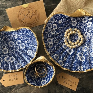 May include: Three decorative ceramic dishes shaped like seashells with a blue and white floral pattern and gold trim. The largest dish is labelled "Trinket Dish", the medium dish is labelled "Ring Dish", and the smallest dish is empty.