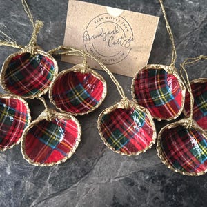 May include: A collection of handmade Christmas ornaments. Each ornament is a shell shape, lined with red, green, and yellow plaid fabric, and trimmed with gold. The ornaments are suspended by gold cords. A small card with text is in the background.