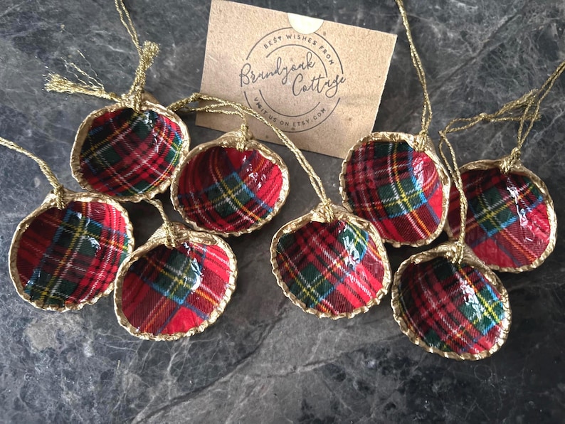 May include: A collection of handmade Christmas ornaments. Each ornament is a shell shape, lined with red, green, and yellow plaid fabric, and trimmed with gold. The ornaments are suspended by gold cords. A small card with text is in the background.