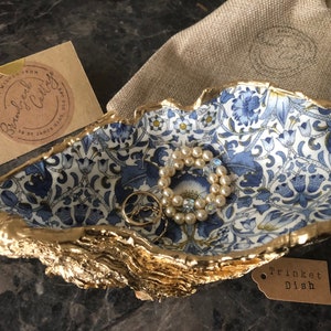 May include: A gold-rimmed trinket dish with a blue and white floral pattern. The dish is shaped like a seashell and has a tag that says "Trinket Dish".