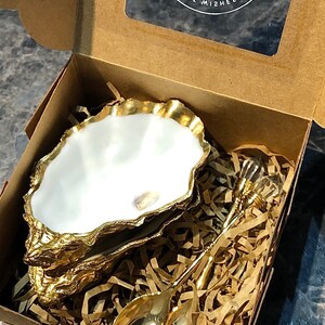 Op de afbeelding: Een gouden oesterschelp-schaal met een wit interieur en een bijpassende gouden lepel. De schaal en de lepel bevinden zich in een bruine kartonnen doos met gescheurd papier.