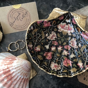 Könnte beinhalten: Eine Muschel-Schmuckschale mit floralem Muster im Inneren und goldfarbenen Rändern. Die Schale ist von anderen goldfarbenen Muscheln, einem kleinen Anhänger mit der Aufschrift "Trinket Dish" und zwei goldenen Ringen umgeben.