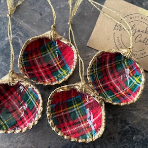 May include: Four seashell ornaments with red, green, and yellow plaid fabric interiors and gold trim. Each ornament has a gold string for hanging. A small card with text is visible in the background.