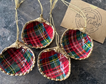 Könnte beinhalten: Vier Muschelschmuckstücke mit rot, grün und gelb karierten Stoffinnenräumen und goldenem Rand. Jedes Ornament hat eine goldene Schnur zum Aufhängen. Im Hintergrund ist eine kleine Karte mit Text zu sehen.