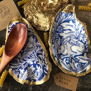 May include: Two gold-leafed oyster shells with blue and white patterned fabric interiors. One shell has a bird design and the other has a rabbit design. A wooden spoon rests in the shell with the bird design. The text "Spoon Rest" is printed on a brown tag.