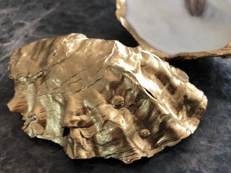 Op de afbeelding: Een gouden geverfde oesterschelp met een getextureerd oppervlak. De schelp heeft een natuurlijke, organische vorm en is perfect om kleine voorwerpen te presenteren.
