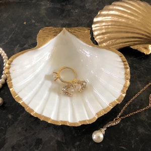 Puede incluir: Un plato de cer&aacute;mica blanca en forma de concha de mar con detalles dorados, que incluye un anillo, dos peque&ntilde;os pendientes con joyas y un collar de perlas con un colgante de perla. El plato est&aacute; sobre una superficie gris oscura.