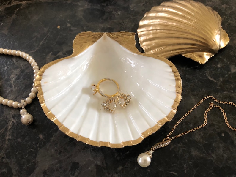 Puede incluir: Un plato de cer&aacute;mica blanca en forma de concha de mar con detalles dorados, que incluye un anillo, dos peque&ntilde;os pendientes con joyas y un collar de perlas con un colgante de perla. El plato est&aacute; sobre una superficie gris oscura.