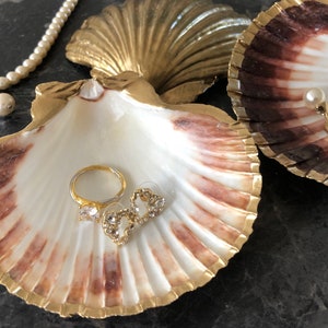 May include: Two seashell dishes with gold accents. One dish is white with brown accents and the other is brown with gold accents. The white dish has a gold ring, two gold heart-shaped earrings, and a pearl necklace with a pearl pendant.