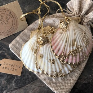 May include: Two white seashells with gold accents and pink highlights are hanging from a burlap bag. The shells are decorated with gold paint and have a delicate, intricate design. The bag has a tag that reads "Hanging Shells".