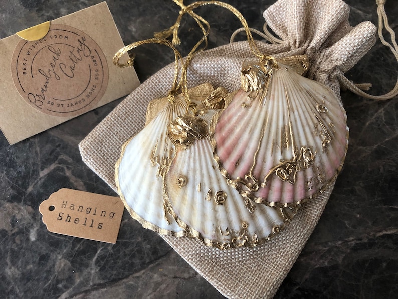 May include: Two white seashells with gold accents and pink highlights are hanging from a burlap bag. The shells are decorated with gold paint and have a delicate, intricate design. The bag has a tag that reads "Hanging Shells".