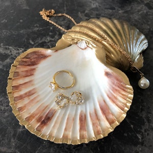 Puede incluir: Una concha de mar pintada de oro con un interior blanco, utilizada como plato para joyas. Contiene un anillo de oro con una piedra transparente, un par de pendientes de oro con piedras transparentes y una cadena de oro con un colgante de perla.