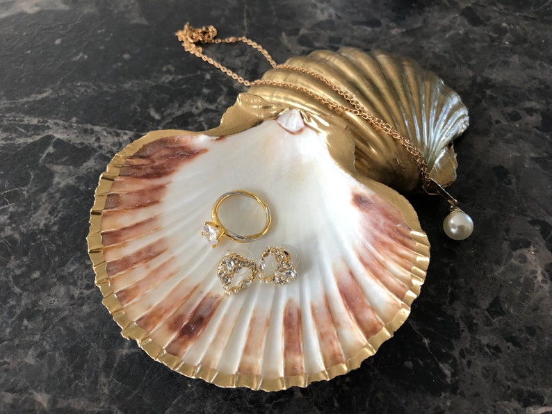 Puede incluir: Una concha de mar pintada de oro con un interior blanco, utilizada como plato para joyas. Contiene un anillo de oro con una piedra transparente, un par de pendientes de oro con piedras transparentes y una cadena de oro con un colgante de perla.