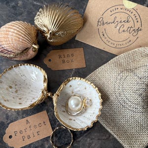 Puede incluir: Cajas de anillos con forma de concha y una peque&ntilde;a bolsa de yute. Una concha abierta muestra un anillo con una perla y una piedra transparente. Las palabras "Ring Box" y "A Perfect Pair" est&aacute;n en peque&ntilde;as etiquetas.