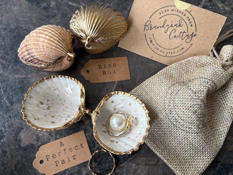 Puede incluir: Cajas de anillos con forma de concha y una peque&ntilde;a bolsa de yute. Una concha abierta muestra un anillo con una perla y una piedra transparente. Las palabras "Ring Box" y "A Perfect Pair" est&aacute;n en peque&ntilde;as etiquetas.